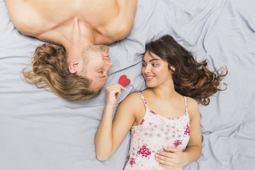 smiling-young-woman-holding-heart-shape-paper-lying-with-her-boyfriend-bed 情侶互動